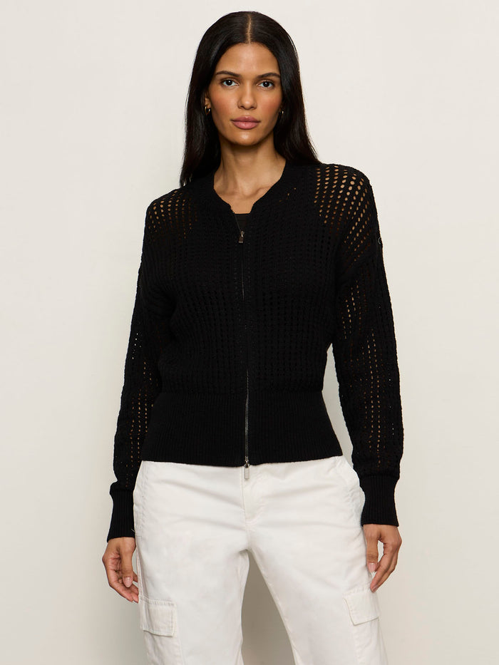 A woman with long dark hair wears the Sanctuary Clothing laguna openwork bomber in black, styled with white pants, standing against a plain light background.