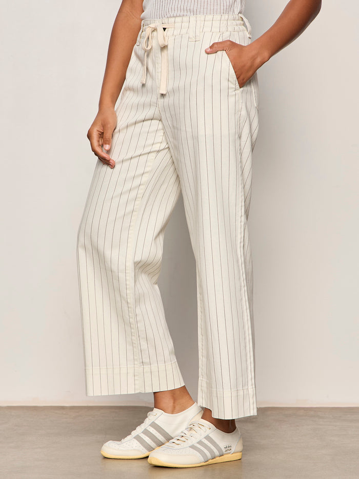 A person wears Sanctuary Clothing's lily cropped pant in setting sun stripe (petite sizing), featuring beige wide-leg, high-waisted, drawstring pants with thin vertical stripes, paired with white sneakers detailed in beige.