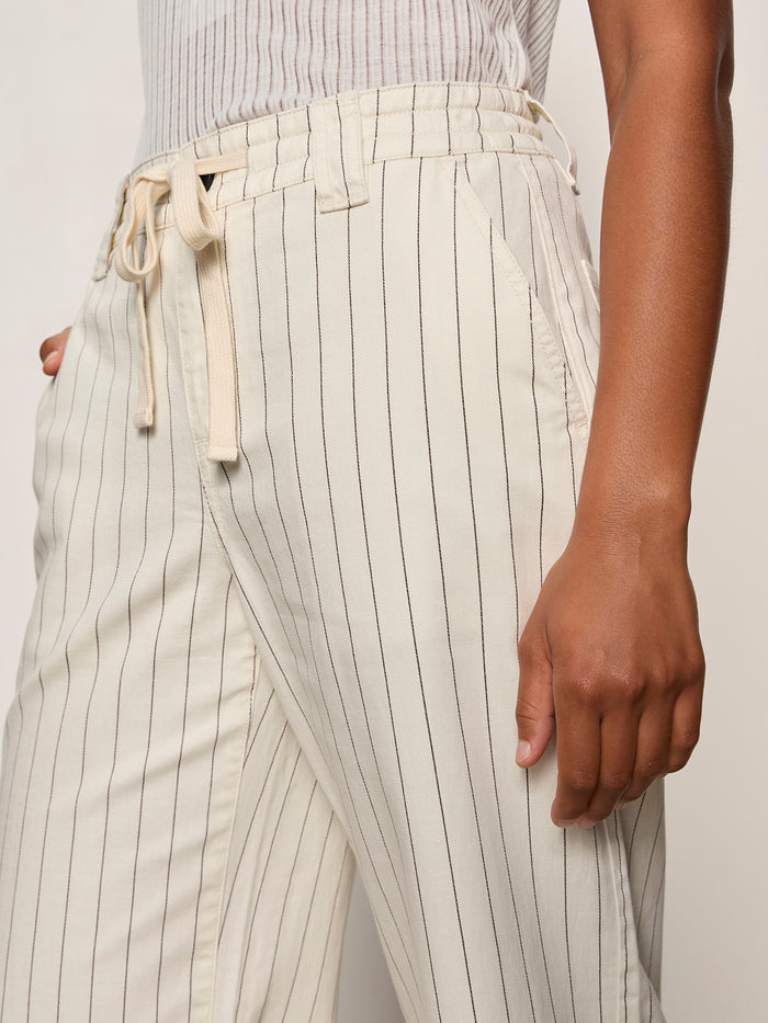 A person wears the Sanctuary Clothing lily cropped pant in setting sun stripe (petite sizing), featuring a cream color, high waist, thin vertical black stripes, drawstring waist, and pockets.