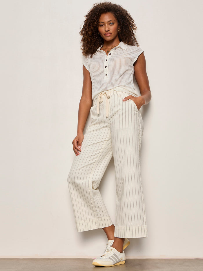 A woman with curly hair wears Sanctuary Clothing's lily cropped pant in setting sun stripe (petite sizing), paired with a short-sleeve button-up shirt and sneakers. She stands against a plain background.