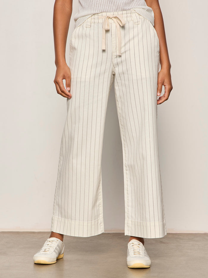 Someone wearing the Sanctuary Clothing lily cropped pant in setting sun stripe—cream-colored with thin vertical stripes and a drawstring waist—paired with white sneakers, stands against a plain background.