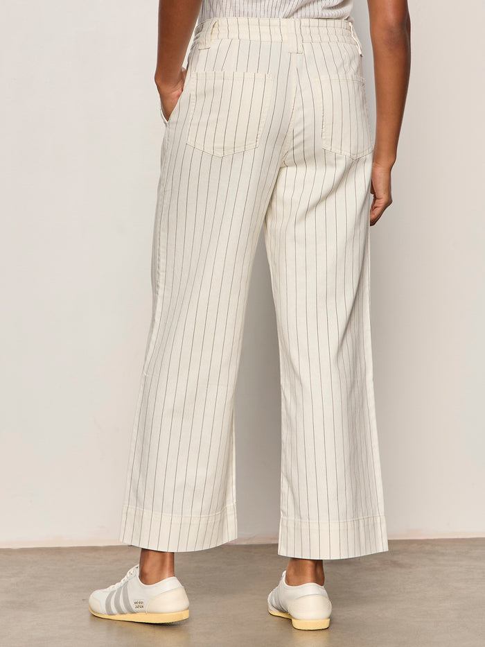A person’s lower half is shown indoors, wearing Sanctuary Clothing’s lily cropped pant in setting sun stripe—a cream wide-leg pant with thin vertical stripes—paired with white sneakers.