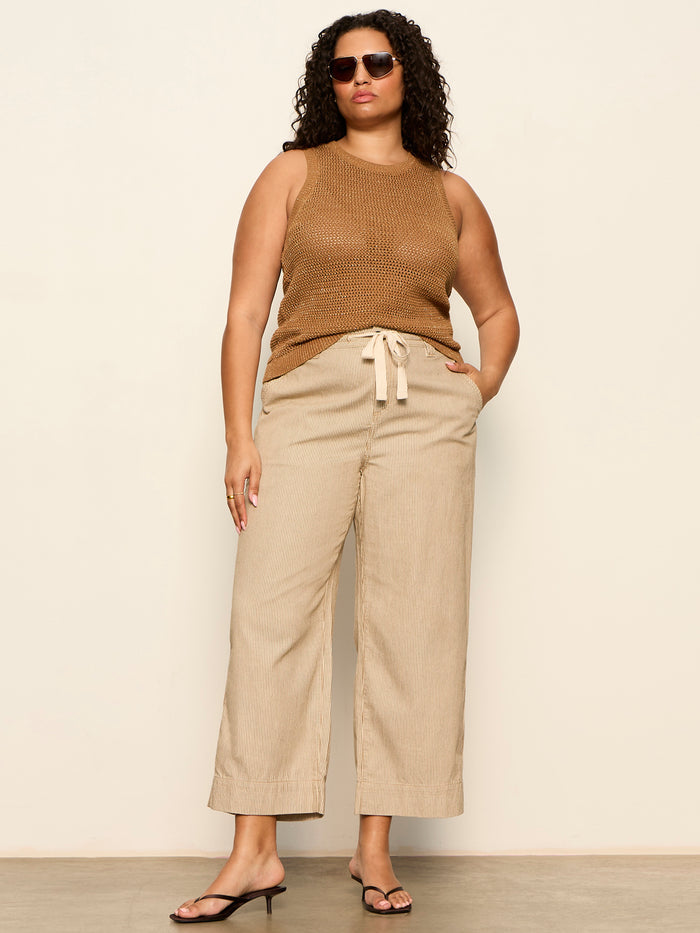 A woman with curly hair and sunglasses wears Sanctuary Clothing's lily stripe cropped pant lido stripe with a sleeveless brown knit top and black heels, posing against a plain background with her hand in her pocket.