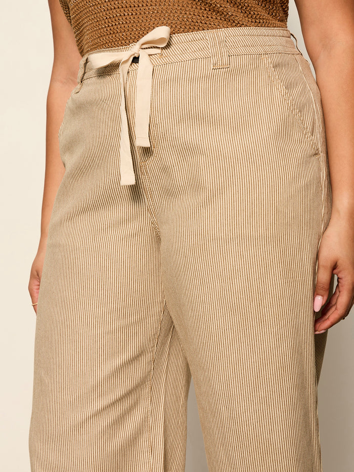 Close-up of a person wearing Sanctuary Clothing's lily stripe cropped pant in lido stripe (extended sizing), featuring light brown vertical stripes and a fabric belt. Only the torso and upper legs are visible; paired with a brown textured top.