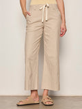 Someone wearing the Sanctuary Clothing lily stripe cropped pant in lido stripe (petite sizing) with a drawstring waist and wide legs, paired with beige sandals, stands against a plain background. One hand is in their pocket.