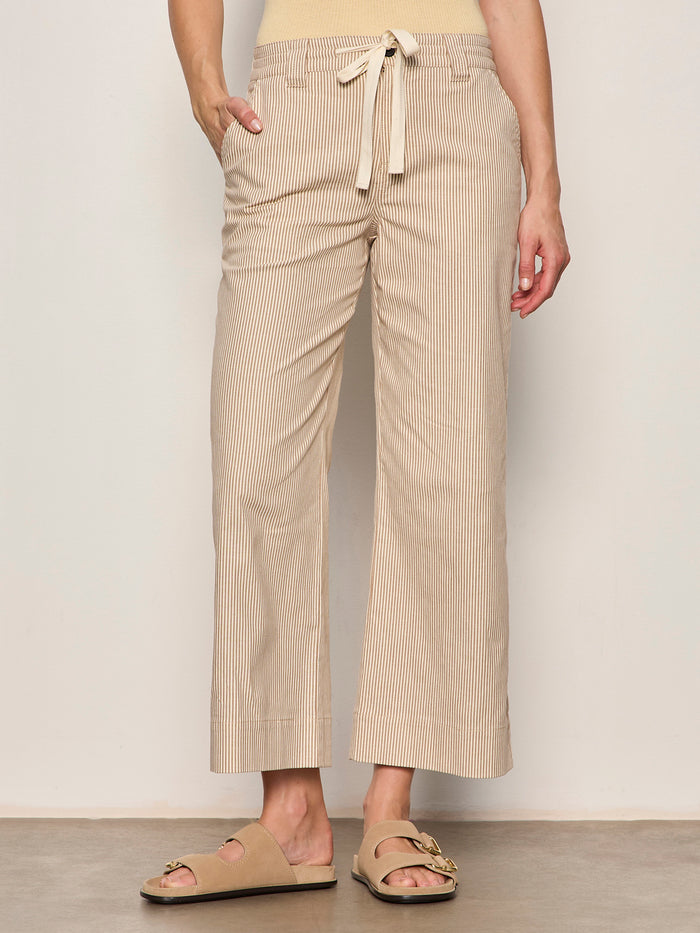 Someone wearing the Sanctuary Clothing lily stripe cropped pant in lido stripe (petite sizing) with a drawstring waist and wide legs, paired with beige sandals, stands against a plain background. One hand is in their pocket.