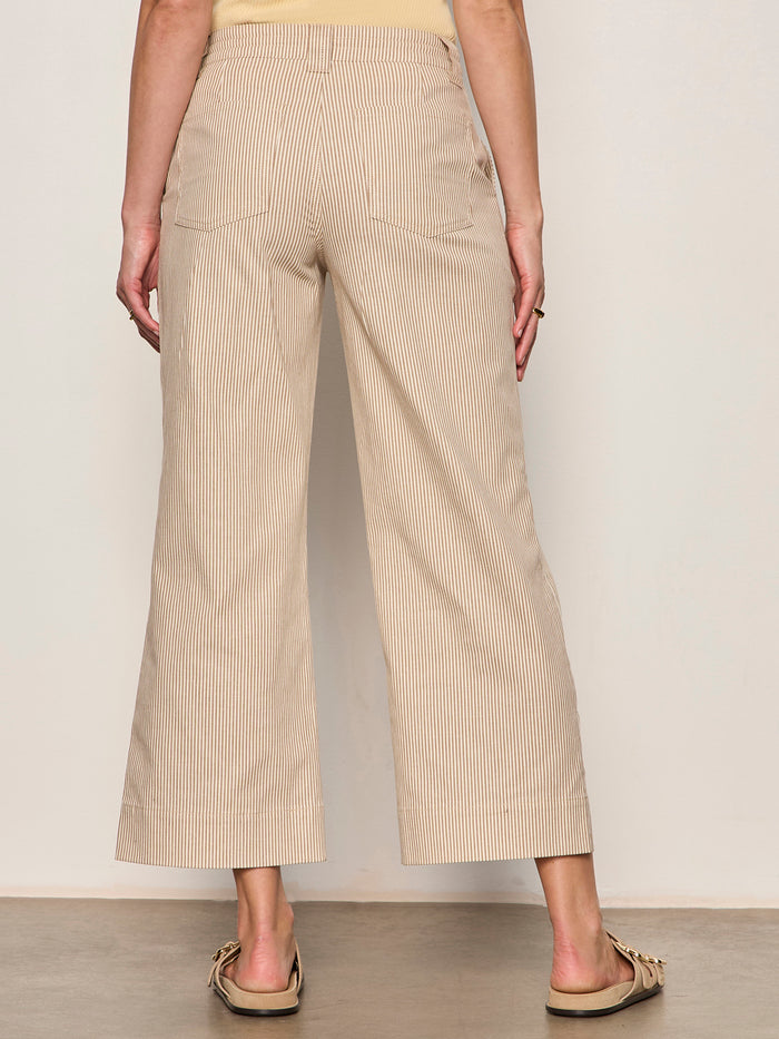 A person stands facing away, wearing Sanctuary Clothing's lily stripe cropped pant in lido stripe petite sizing and beige moccasin-style shoes, with a gray floor and a plain light-colored wall in the background.