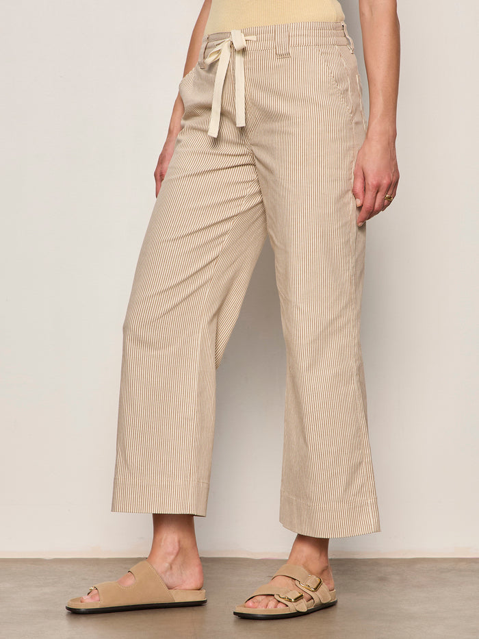 A person wears Sanctuary Clothing's lily stripe cropped pant in lido stripe (petite), paired with a yellow top and tan sandals, standing against a light-colored wall.