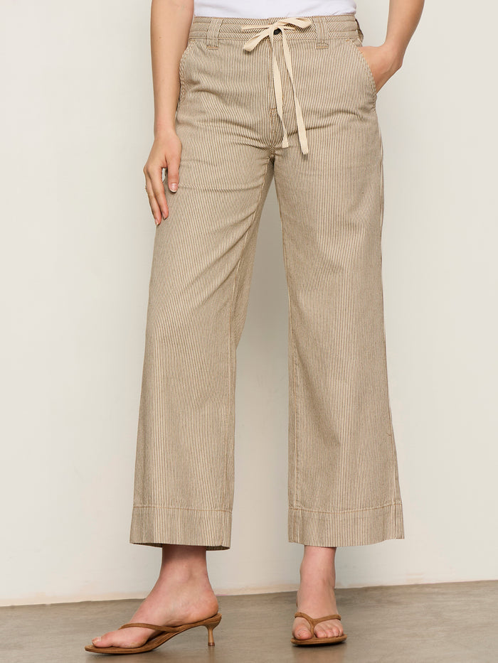 A person wearing Sanctuary Clothing's lily stripe cropped pant lido stripe, featuring beige and white pinstripes and a drawstring waist, paired with a white top and tan heeled sandals against a plain light background.