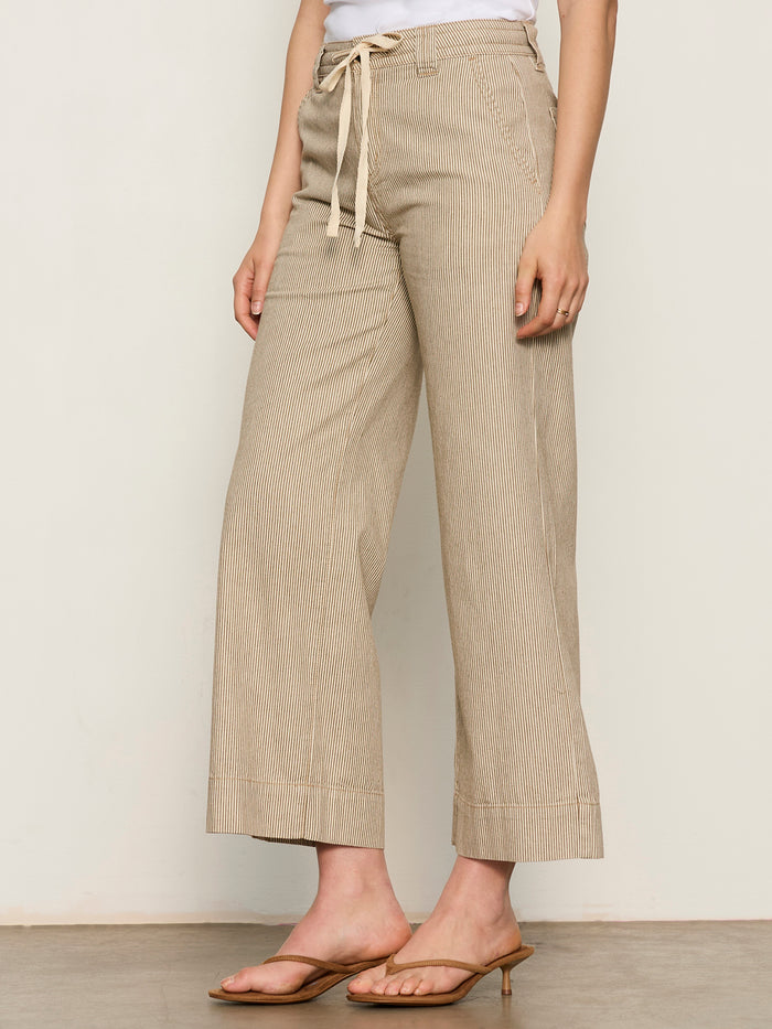 A person wearing Sanctuary Clothing's lily stripe cropped pant in lido stripe, featuring beige wide-leg, high-waisted, drawstring style with subtle stripes, paired with a white top and tan heeled sandals against a neutral background.