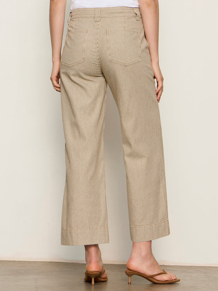 A person stands facing away wearing the Sanctuary Clothing lily stripe cropped pant in lido stripe, paired with a white top and tan heeled sandals, against a plain light-colored background.