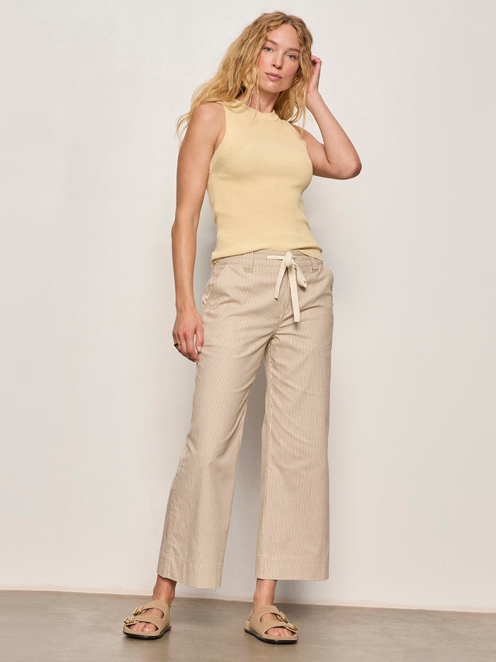 A woman with wavy blonde hair wears Sanctuary Clothing's lily stripe cropped pant in lido stripe, a sleeveless beige top, and beige sandals. She stands against a plain light wall, one hand on her head and the other by her side.