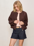 A woman with blonde hair wears the Sanctuary Clothing linen bubble jacket in rich soil over a light pink top, dark denim shorts, and a light pink belt. She looks down, holding the edges of her jacket.