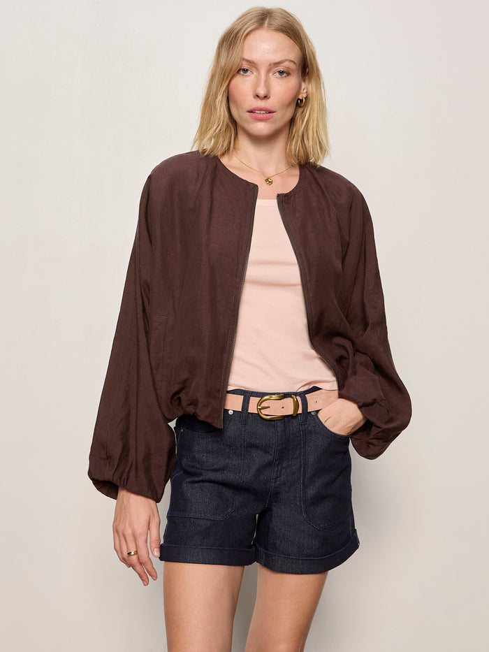 A woman with blonde hair models the Sanctuary Clothing linen bubble jacket in rich soil over a light pink top, dark denim shorts, and a beige belt, standing against a plain light-colored background with one hand in her pocket.