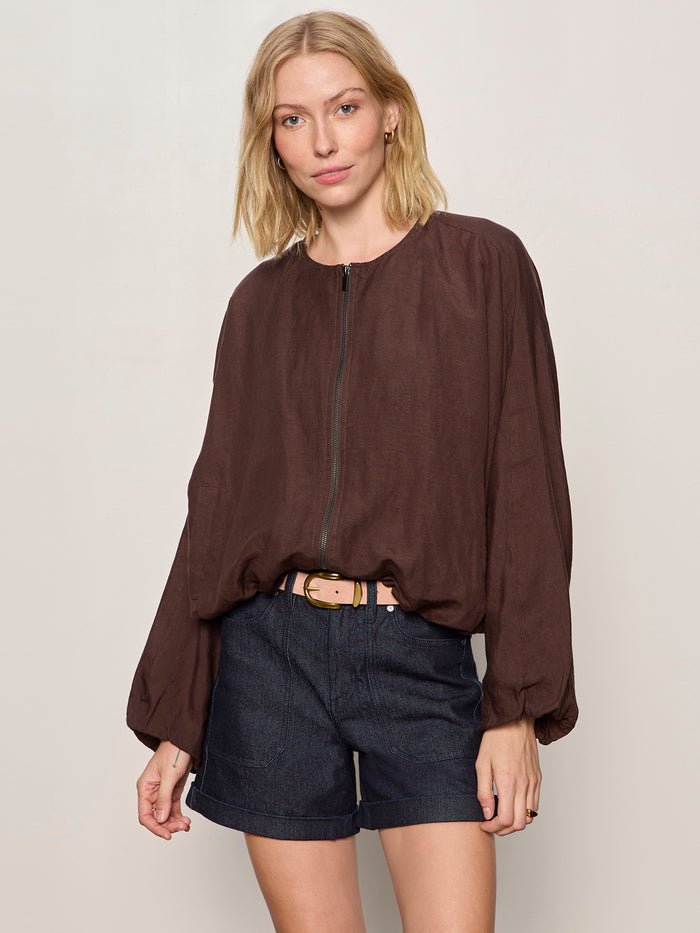 A woman with shoulder-length blonde hair and a neutral expression wears the Sanctuary Clothing linen bubble jacket in rich soil against a plain background, paired with dark denim shorts and a light belt.