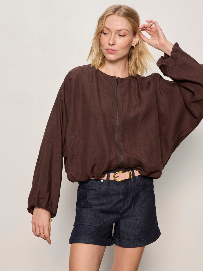 A woman with blonde hair wears the Sanctuary Clothing linen bubble jacket in rich soil and dark denim shorts with a beige belt, standing against a plain light background. She looks to the side, touching her hair with one hand.