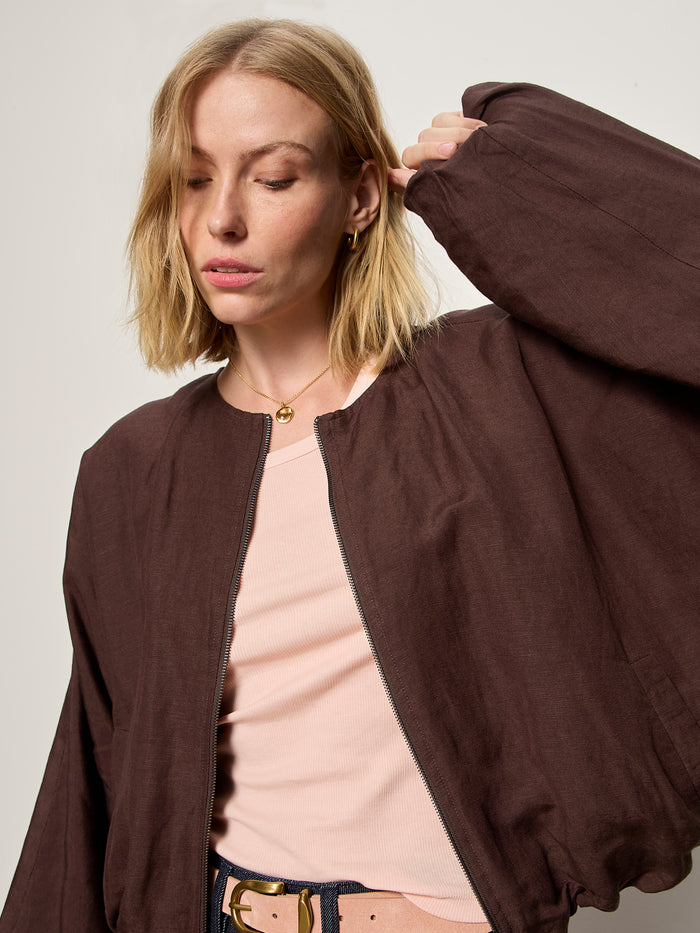 A woman with shoulder-length blonde hair wears Sanctuary Clothing's linen bubble jacket in rich soil over a pale pink top. She stands against a plain light background, eyes closed, one arm raised.