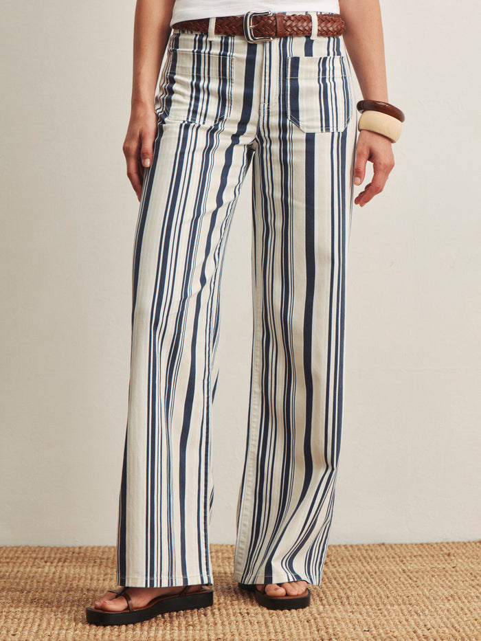 A person wearing the Sanctuary Clothing marine full length sail stripe pants, featuring high-waisted, wide-leg blue and white stripes, styled with a brown woven belt, sandals, and chunky bangles on a textured mat against a plain background.