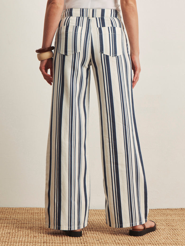 A person stands on a woven rug, wearing Sanctuary Clothing's marine full length sail stripe pants with vertical blue and white stripes, black sandals, and chunky bangles. The photo is taken from behind, showing the back pockets of the pants.