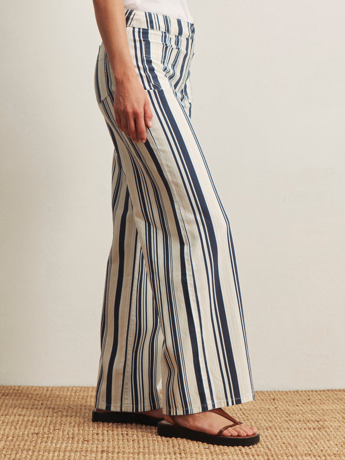 A person stands on a woven rug, wearing Sanctuary Clothing's marine full length sail stripe pants—white with vertical navy blue stripes—paired with black sandals. Only the lower body is shown against a plain light background.