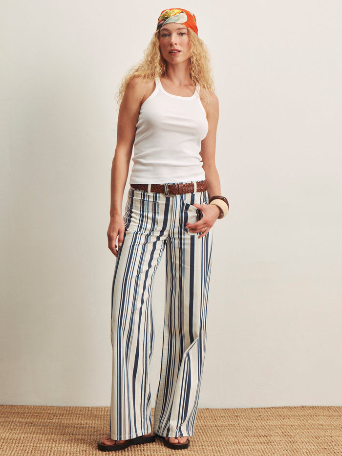 A woman with long, curly blonde hair wears Sanctuary Clothing’s marine full length sail stripe pants, a white tank top, a colorful headscarf, a brown belt, sandals, and bracelets while standing on a woven rug against a plain background.