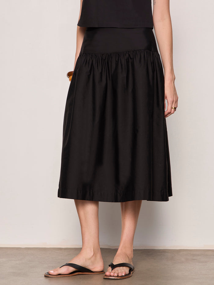 Someone wearing the Sanctuary Clothing "Matter of Time" poplin skirt in black, paired with a black sleeveless top and sandals, stands on a neutral floor against a light background.