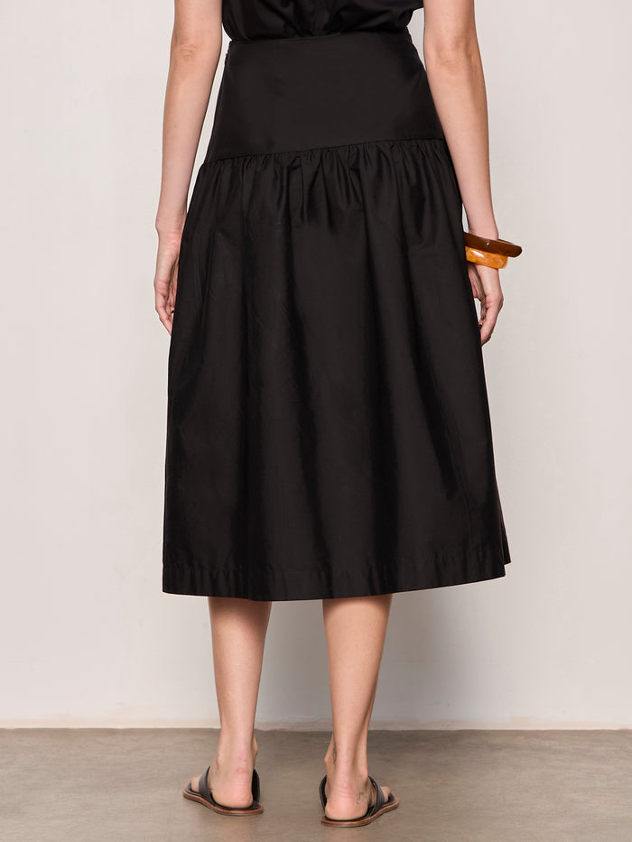 A person stands on a concrete floor wearing the Sanctuary Clothing "matter of time poplin skirt" in black, paired with a brown bracelet and black sandals. Their back is visible from shoulders down, set against a plain light-colored wall.