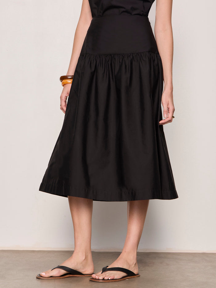 A person wearing the Sanctuary Clothing "matter of time" poplin skirt in black, styled with black sandals, a sleeveless black top, and bracelets on one wrist, stands against a light-colored wall.