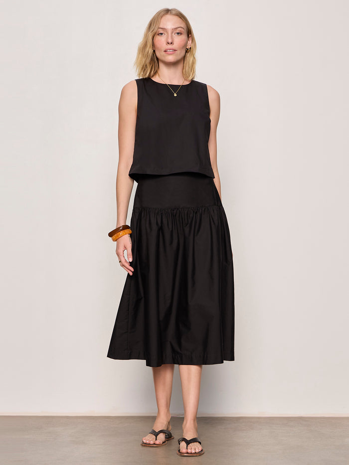 A woman stands against a light background, wearing Sanctuary Clothing’s matter of time poplin skirt in black with a sleeveless black top, necklace, brown bangle, and black sandals.