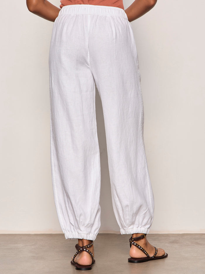 A person is indoors, facing away, wearing a peach sleeveless top and Sanctuary Clothing's melody micro linen balloon white pants with elastic hems, paired with black studded sandals. The background is plain and light-colored.