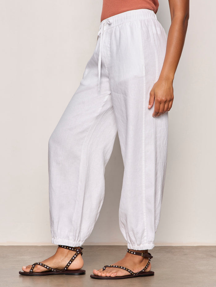 A person wearing Sanctuary Clothing's melody micro linen balloon white pants, featuring an elastic cuff and drawstring waist, stands sideways in studded black sandals against a neutral background.