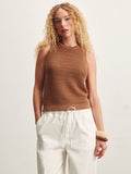 A woman with long curly blonde hair wears the Sanctuary Clothing natural state halter tank in toasted coconut metallic, paired with loose white drawstring pants and chunky bracelets, standing against a plain light background.