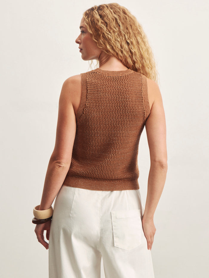 A woman with curly blonde hair wears the Sanctuary Clothing natural state halter tank in toasted coconut metallic and white high-waisted pants, standing with her back to the camera against a plain light background, accessorized with large bangles.
