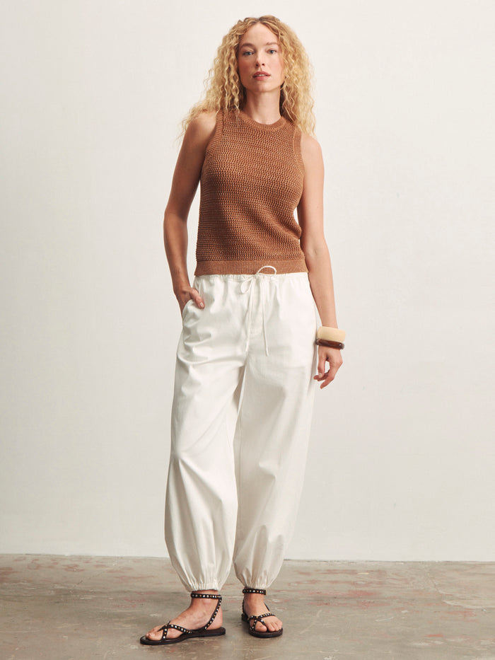 A person with long, curly blond hair wears Sanctuary Clothing's natural state halter tank in toasted coconut metallic, paired with loose white drawstring pants, black sandals, and a large bracelet against a plain off-white background.