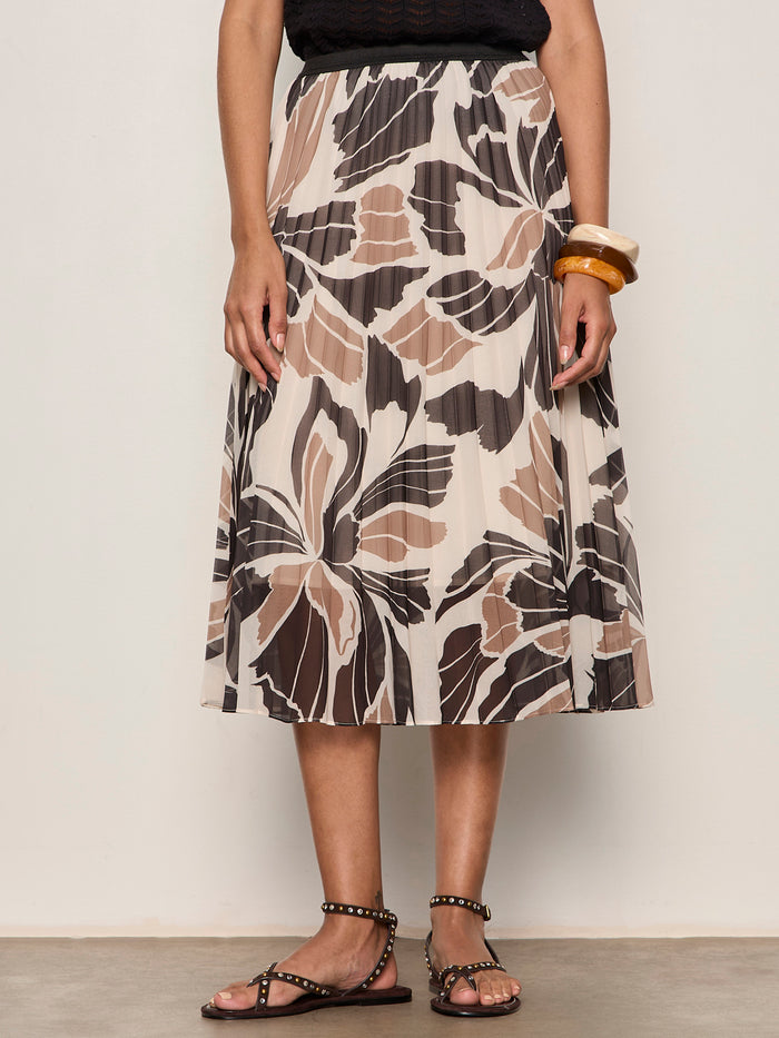 A person wearing a black sleeveless top, the Sanctuary Clothing nostalgia pleated midi skirt lotus with abstract floral patterns, black studded sandals, and chunky bangles on one wrist.