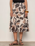 A person wearing a black sleeveless top, the Sanctuary Clothing nostalgia pleated midi skirt lotus with abstract floral patterns, black studded sandals, and chunky bangles on one wrist.