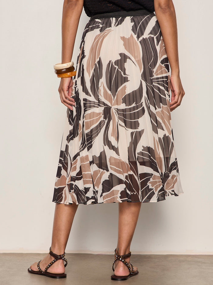 Wearing the Sanctuary Clothing nostalgia pleated midi skirt lotus, a person in a black top, studded strappy sandals, and chunky bangles stands with their back to the camera against a plain background.