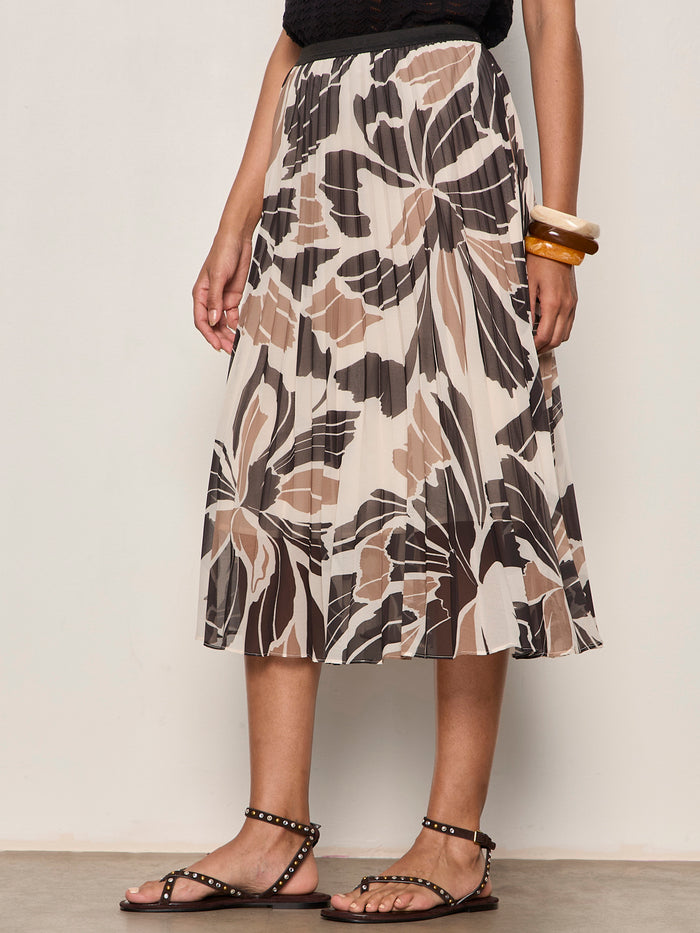 A person wears the Sanctuary Clothing nostalgia pleated midi skirt lotus—a pleated, abstract floral midi—paired with a black sleeveless top, studded black sandals, and chunky bangles.