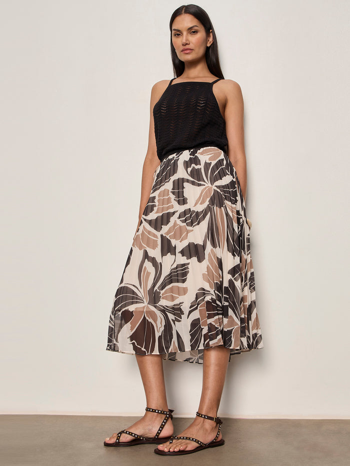 A woman stands against a plain wall wearing a sleeveless black top, black studded flat sandals, and the Sanctuary Clothing nostalgia pleated midi skirt lotus featuring a large neutral floral print.