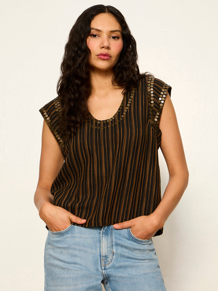 A woman with long, wavy dark hair wears the Sanctuary Clothing outsider embellished shell top in dark cumin black stripe, featuring gold vertical stripes and shoulder details. She stands against a plain light background with one hand in her jeans pocket.