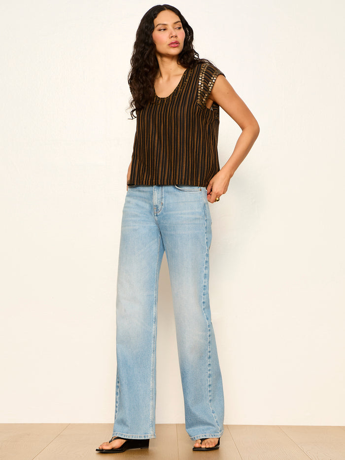 A woman with long dark hair wears the Sanctuary Clothing outsider embellished shell top in dark cumin black stripe, paired with light blue wide-leg jeans and black sandals, standing against a plain white wall.