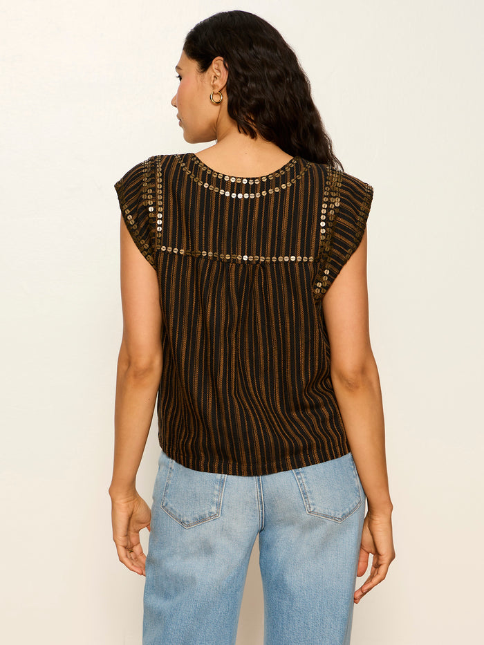 A woman with wavy brown hair wears Sanctuary Clothing's Outsider Embellished Shell Top in dark cumin black stripe, paired with light blue jeans. She stands with her back to the camera against a plain light wall.