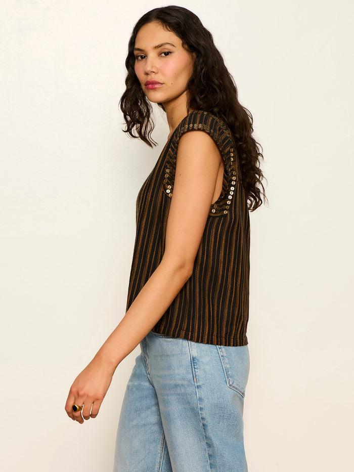 A woman with long curly hair models the Sanctuary Clothing outsider embellished shell top in dark cumin black stripe with decorative shoulder buttons and light blue jeans, standing sideways against a plain off-white background.