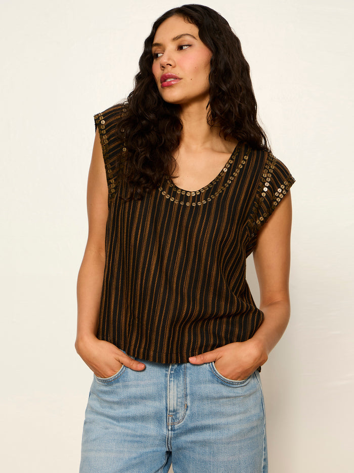 A woman with long wavy hair wears the Sanctuary Clothing Outsider Embellished Shell Top in dark cumin black stripe and light blue jeans, posing against an off-white background with her hands in her pockets.