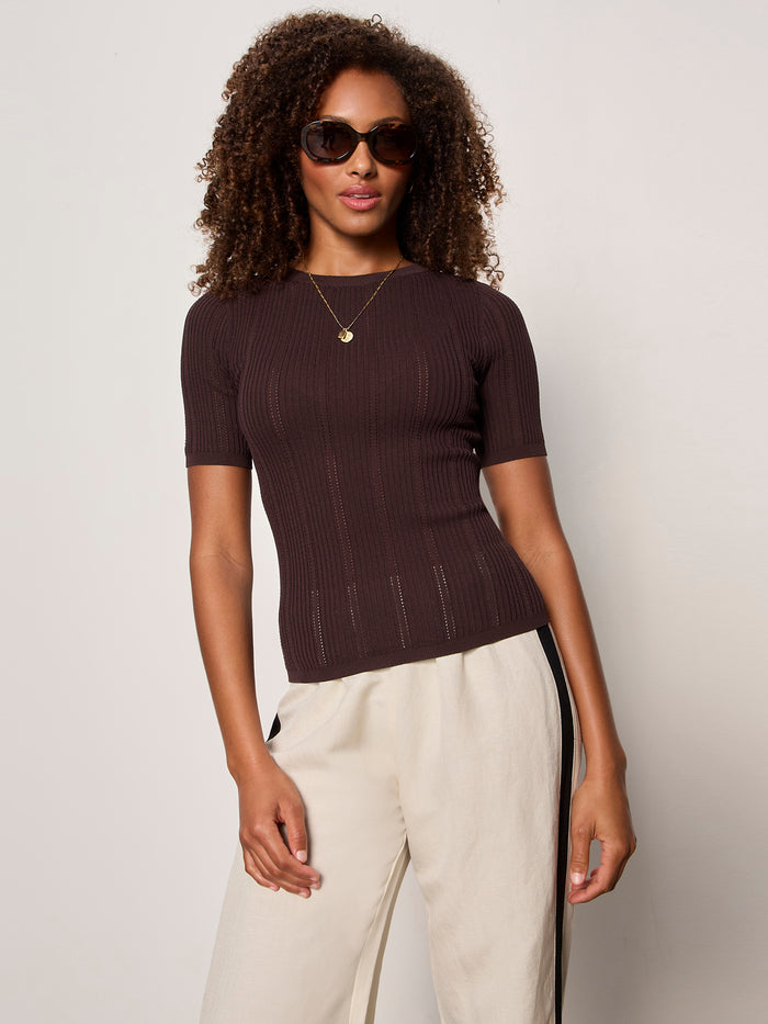 A woman with curly hair poses against a plain light background, wearing Sanctuary Clothing's perfect knitwear tee in rich soil and cream pants with black side stripes, styled with sunglasses.