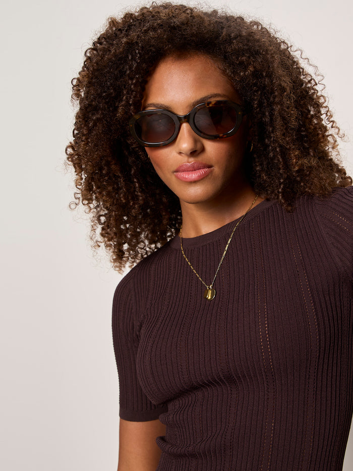 A woman with curly hair wears large dark sunglasses, a gold necklace, and the Sanctuary Clothing perfect knitwear tee in rich soil, standing against a light background.