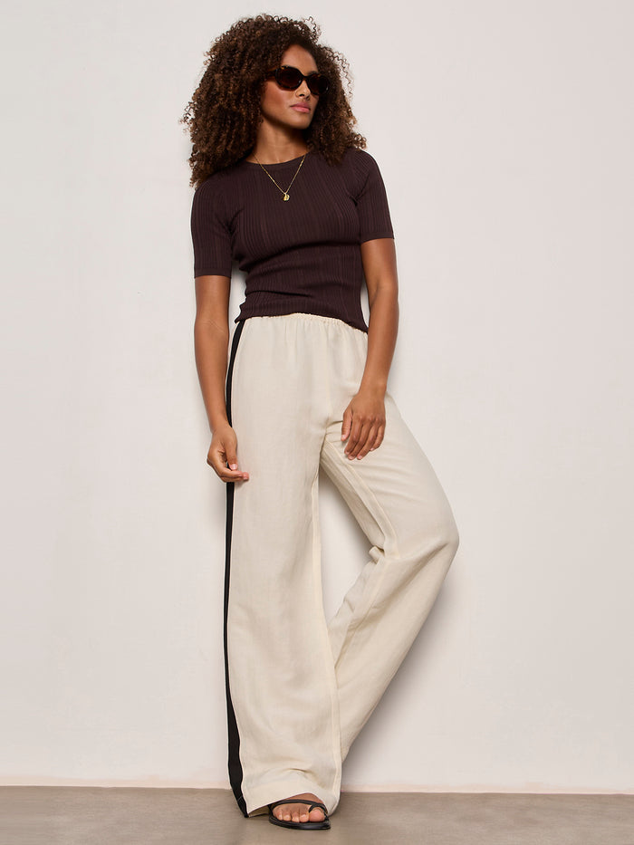 A woman with curly hair and sunglasses wears the Sanctuary Clothing "perfect knitwear tee rich soil," paired with loose cream wide-leg pants featuring black side stripes, sandals, and layered gold necklaces against a plain light background.