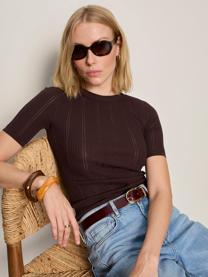 A woman with blonde hair and sunglasses sits confidently on a woven chair, wearing the Sanctuary Clothing perfect knitwear tee in rich soil and blue jeans, accessorized with large bangles, against a neutral background.