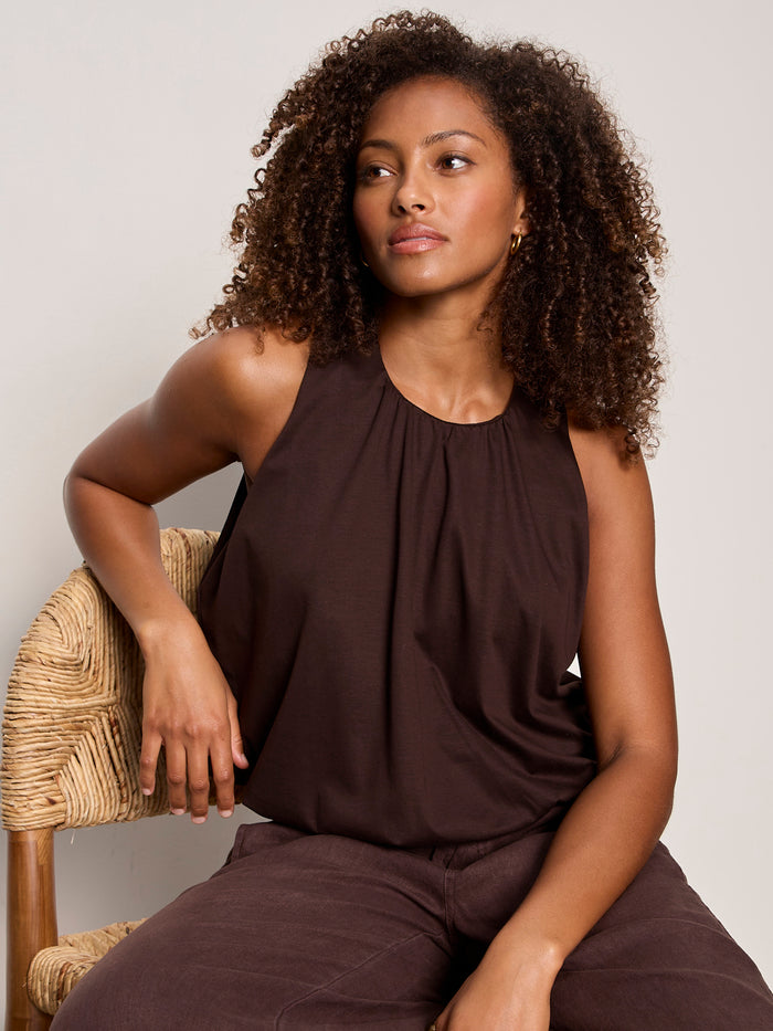 A woman with curly hair sits on a woven chair, wearing the Sanctuary Clothing retreat bubble tank in rich soil, paired with matching pants, gazing calmly to the side against a neutral background.
