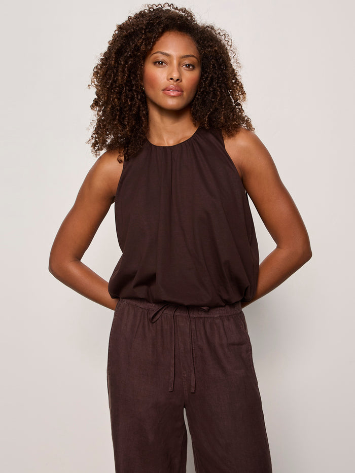 A woman with curly hair wears the Sanctuary Clothing retreat bubble tank in rich soil, a sleeveless, loose-fitting dark brown top, standing against a plain light background with her hands relaxed at her sides.